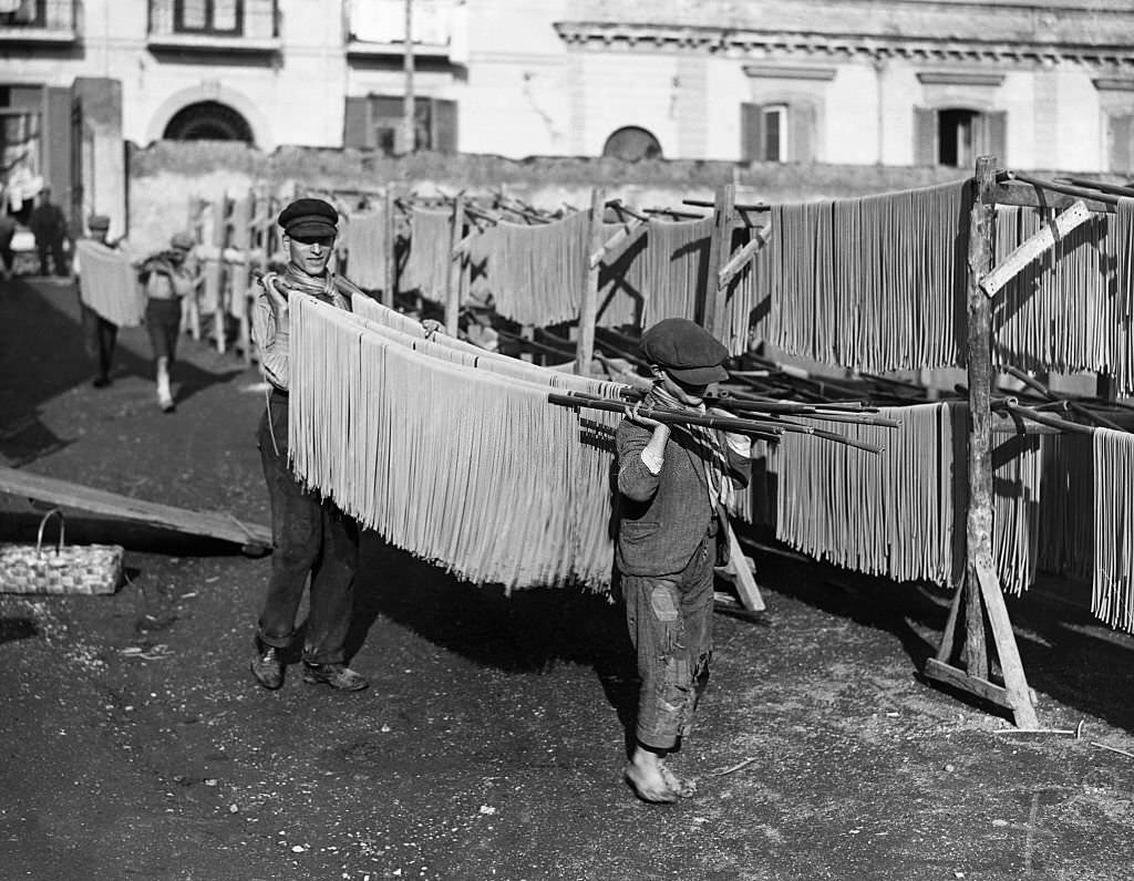 Young boys carry poles of spaghetti into a factory yard for drying, 1920s