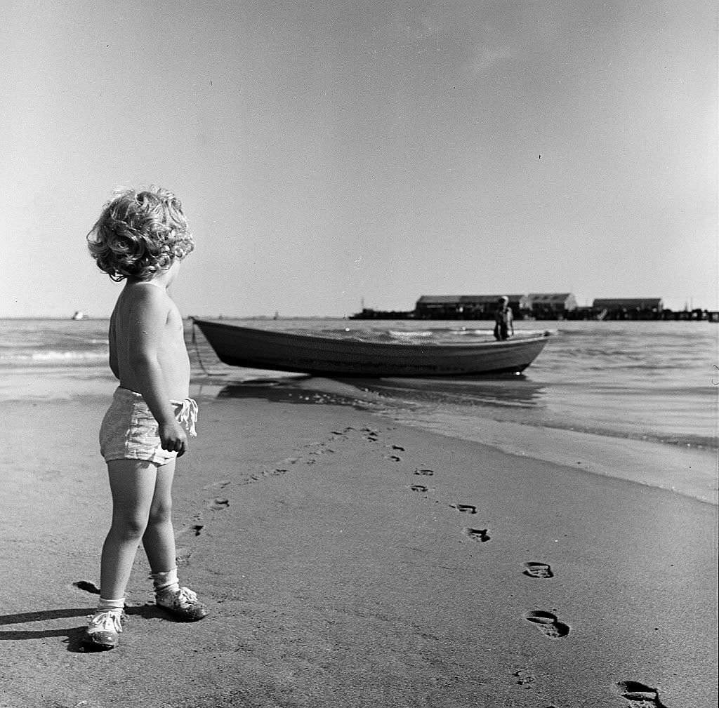 A young child looks back towards an unidentified figure standing next to a boat while on the beach, Provincetown, Massachusetts, 1948.