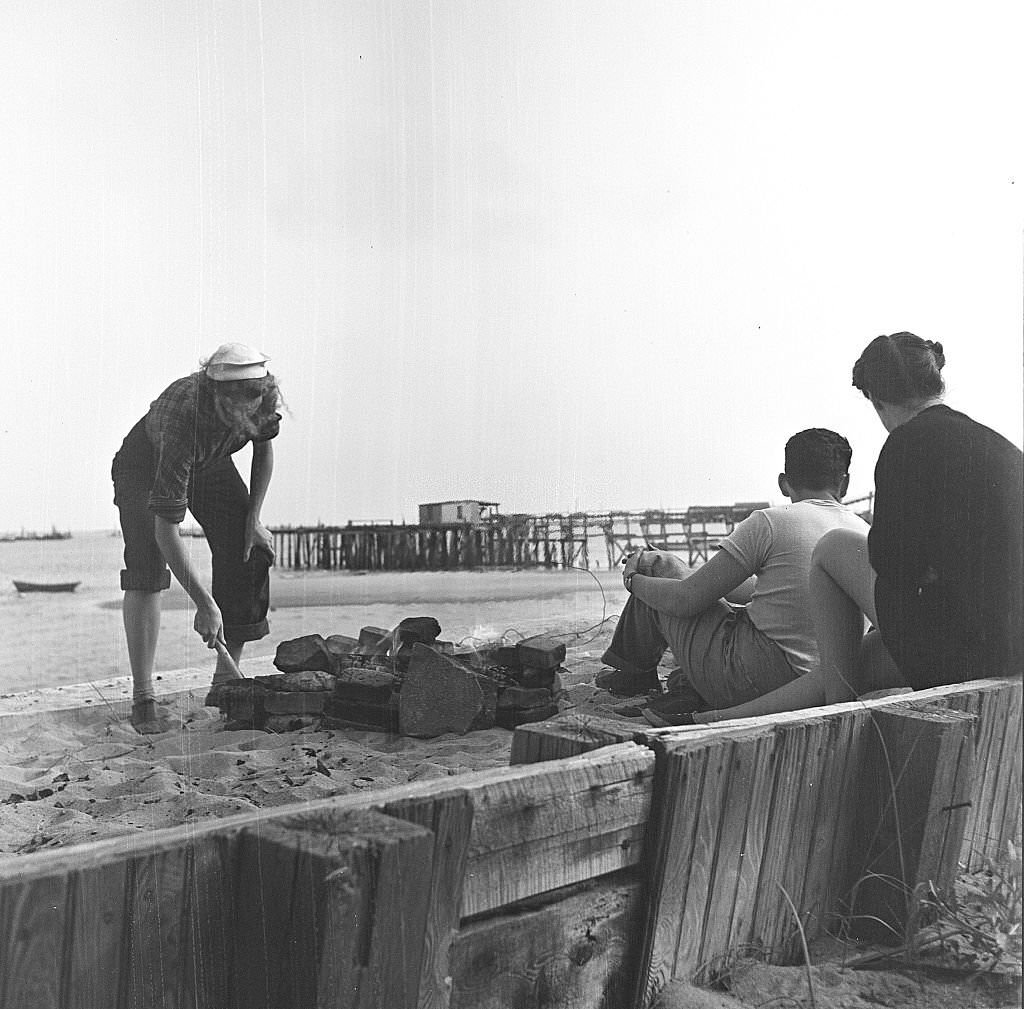 A man and a woman sit while another woman tends the fire for their beachside cookout, Provincetown, Massachusetts, 1948.