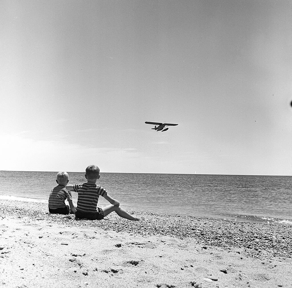 Two young boys sit on the beach and watch as a seaplane lands on Cape Cod, Provincetown, Massachussetts, 1947.