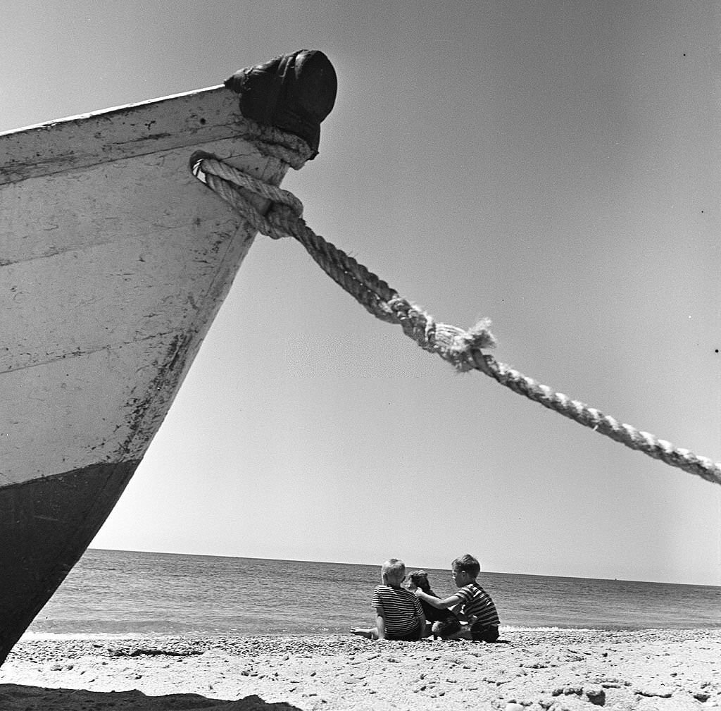 Two young boys sit on the beach with their dog, with a rowboat anchored in the foreground on Cape Cod, Provincetown, Massachussetts, 1947.