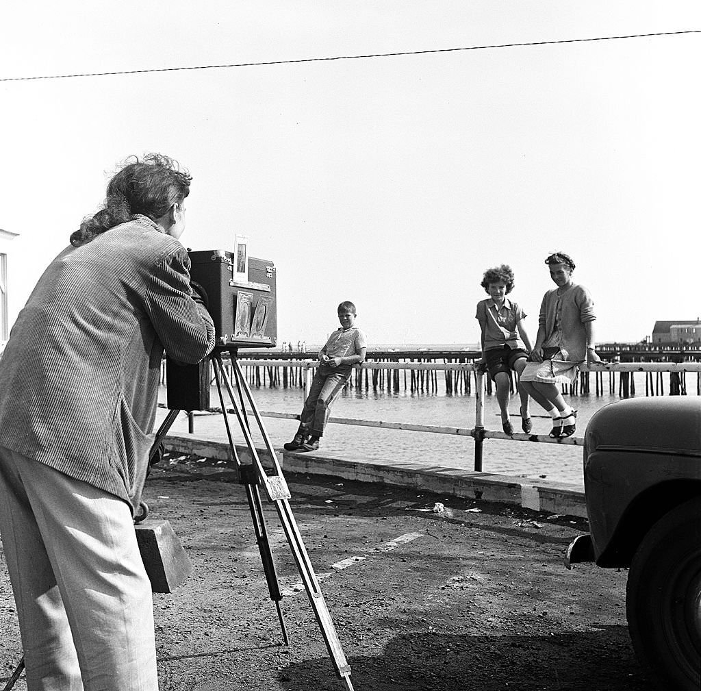 hree visitors sit or lean on the pier's railings as a photographer uses a camera on a tripod to take portraits, Provincetown, Massachusetts, 1948.