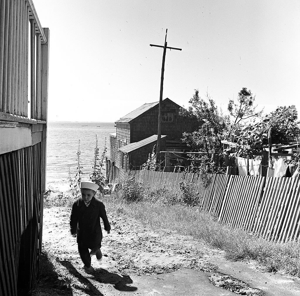 A young boy wearing a sailor's hat walks past the deck of a seaside house on Cape Cod, Provincetown, Massachussetts, 1947.