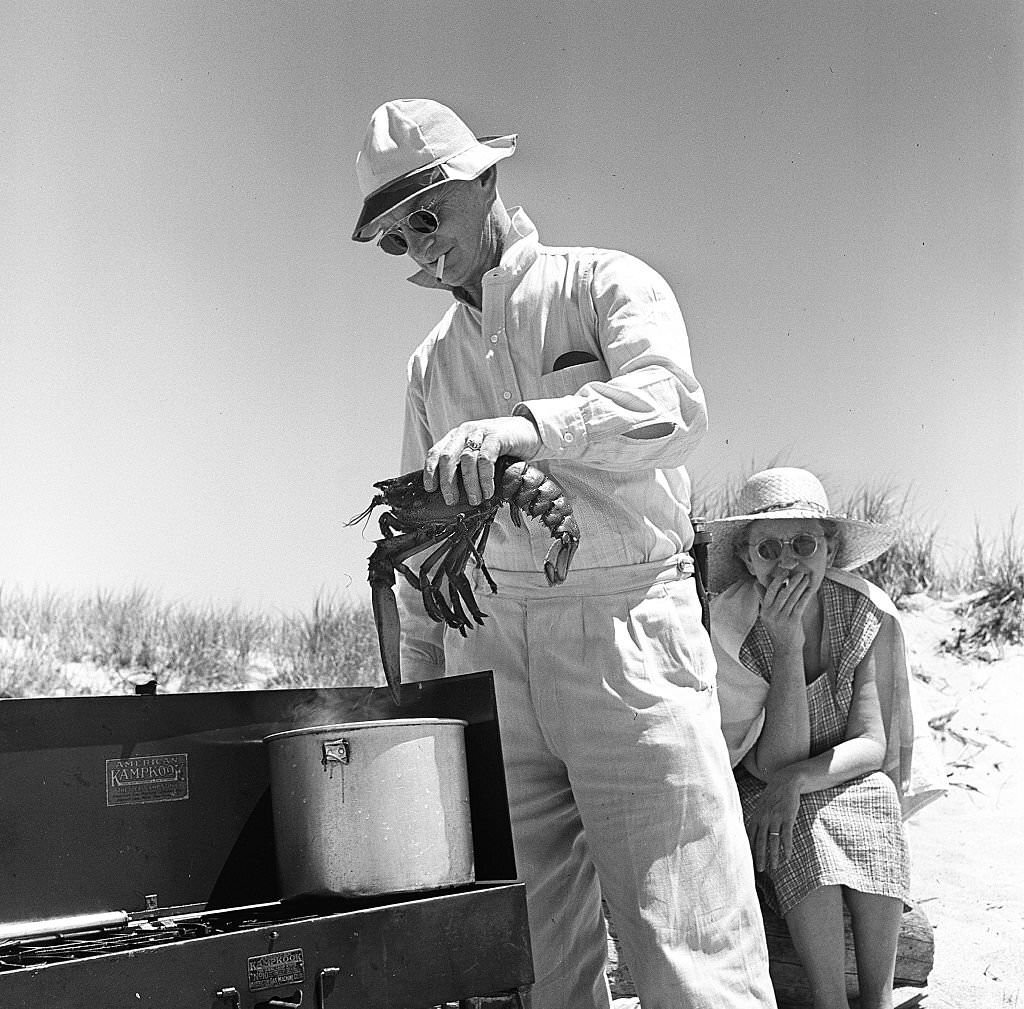 A man prepares to cook a lobster in a pot set up on an outdoor grill, with a woman seated behind him on Cape Cod, Provincetown, Massachussetts, 1947.