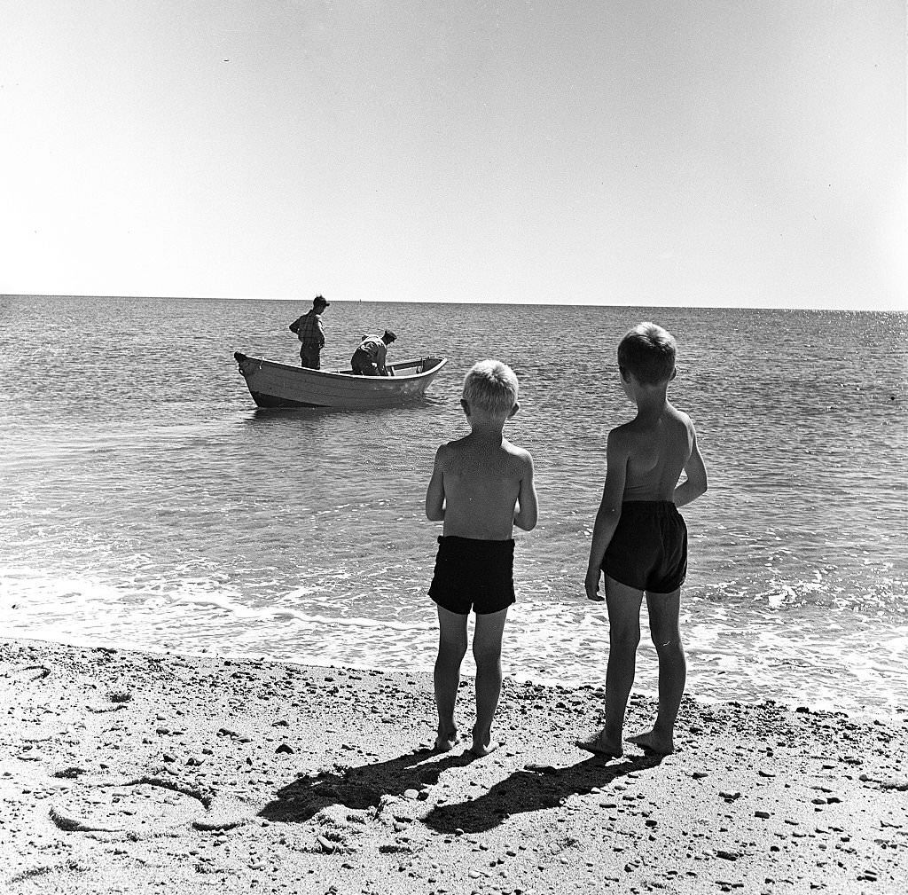 Two young boys watch as two fishermen head out on a small boat on Cape Cod, Provincetown, Massachussetts, 1947.