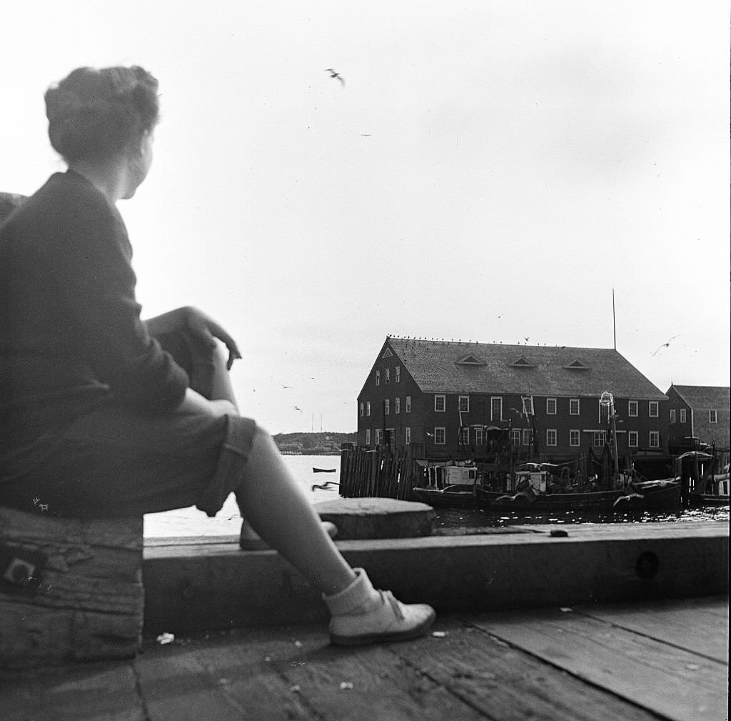 A young woman sitting on the pier looks out onto some dockside buildings on Cape Cod, Provincetown, Massachussetts, 1947.