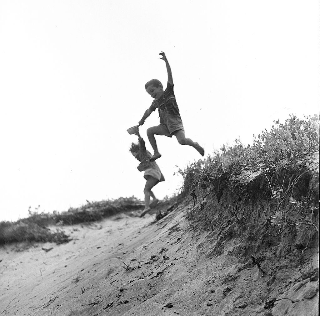 Two young children, a boy and a girl, hold hands as they jump and play in the sand dunes of the beach, Provincetown, Massachusetts, 1948.