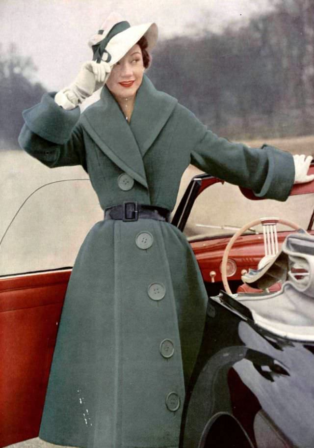 Sophie Malgat in green wool coat with giant buttons lined diagonally, waist is cinched by patent leather belt, worn with white piqué hat, by Jacques Fath, 1951
