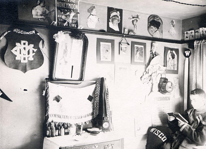 Man sitting in boarding house or fraternity house room reading. Pinups and other decorations on south wall, 1912.