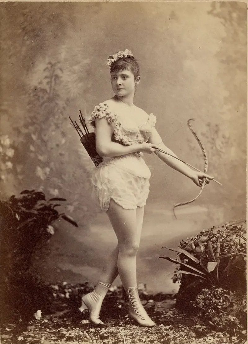 Miss Farrington in a short sleeveless costume, holding bow and arrow, with quiver and arrows on back, buttoned and heeled boots.