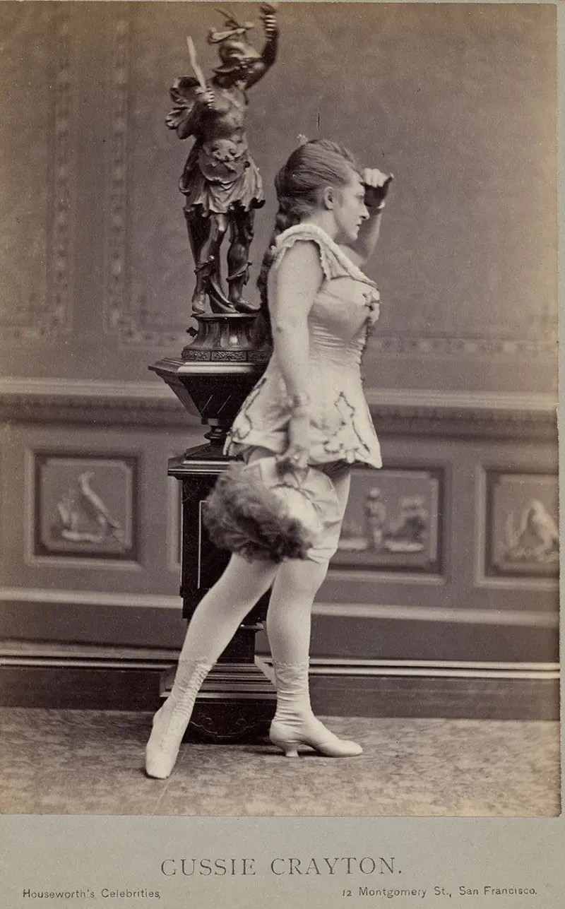Gussie Crayton in hat with feather, short sleeveless costume with half-calf laced boots.