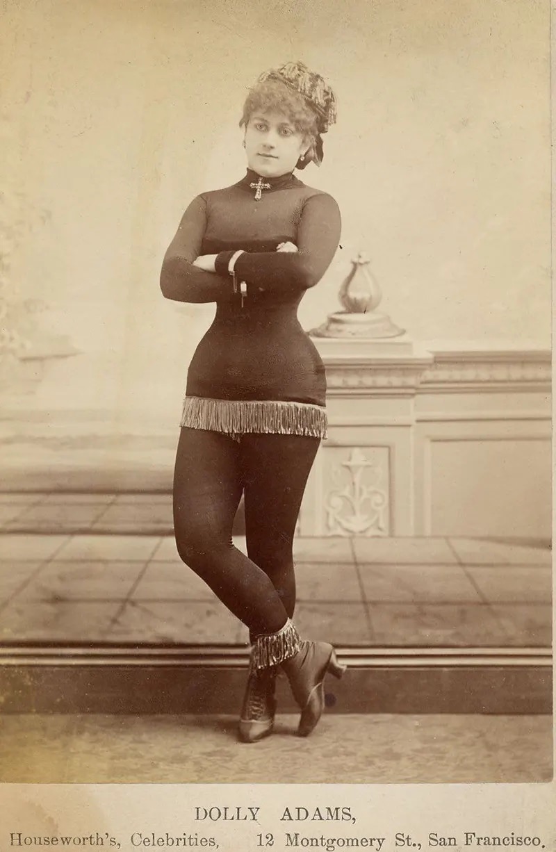 Dolly Adams with fringe at the bottom of a short costume, tights, short-heeled boots topped with fringe, cross at neck, cap.