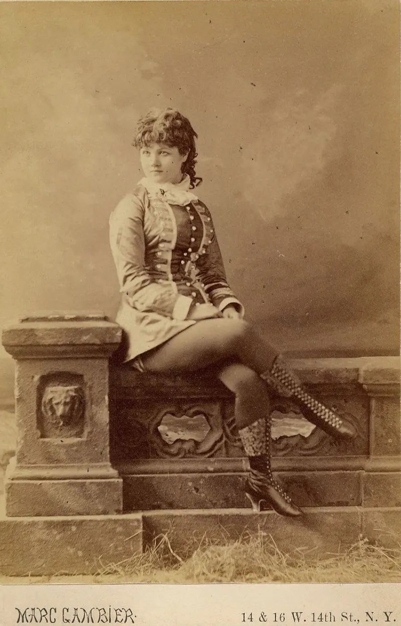 Minnie Marshall sitting on a stone fence, in a short dress and boots.