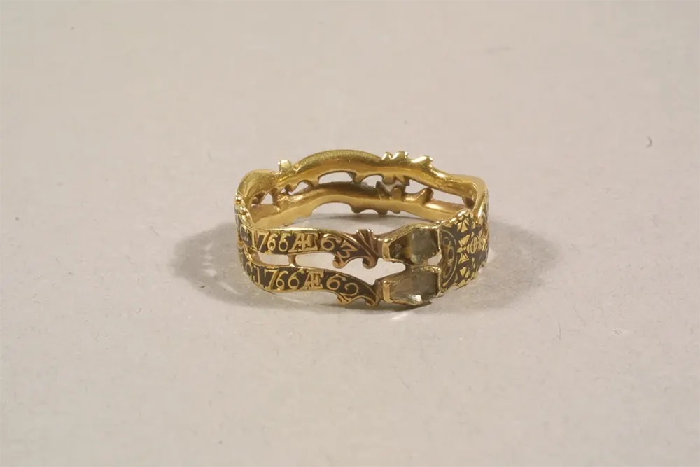 A gold mourning ring with black enamel, featuring two coffin-shaped bezels with faintly visible skeletons.