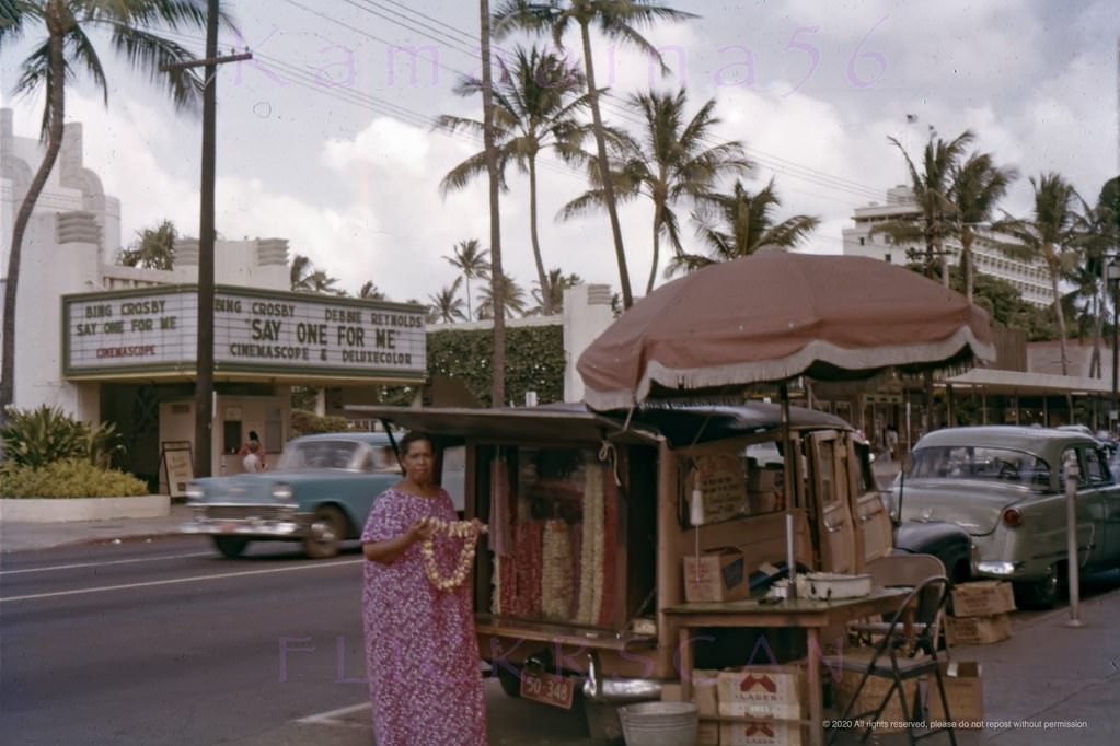 Waikiki’s Kalakaua Avenue viewed from the makai (ocean) side of the street with a sidewalk lei seller operating out of an old woodie in the foreground, 1959