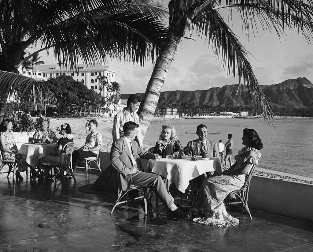 Couples sit outdoors at patio tables, having a meal at a beach side restaurant while a waiter looks on, Waikiki Beach, 1945