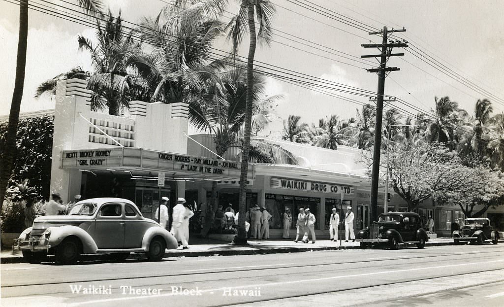 Sailors mill outside of Waikiki theater block on quiet street with parked cars and palm trees, 1940s