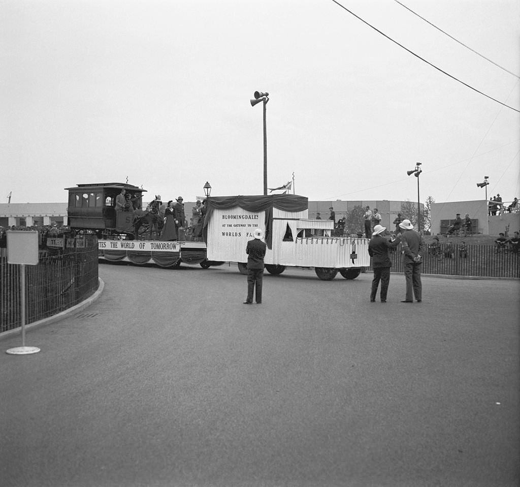 Crowds of people and security staff, watching a carnival float carrying a western type horsedrawn tram and people dressed in 1890's period costumes, drawn by a motor vehicle.