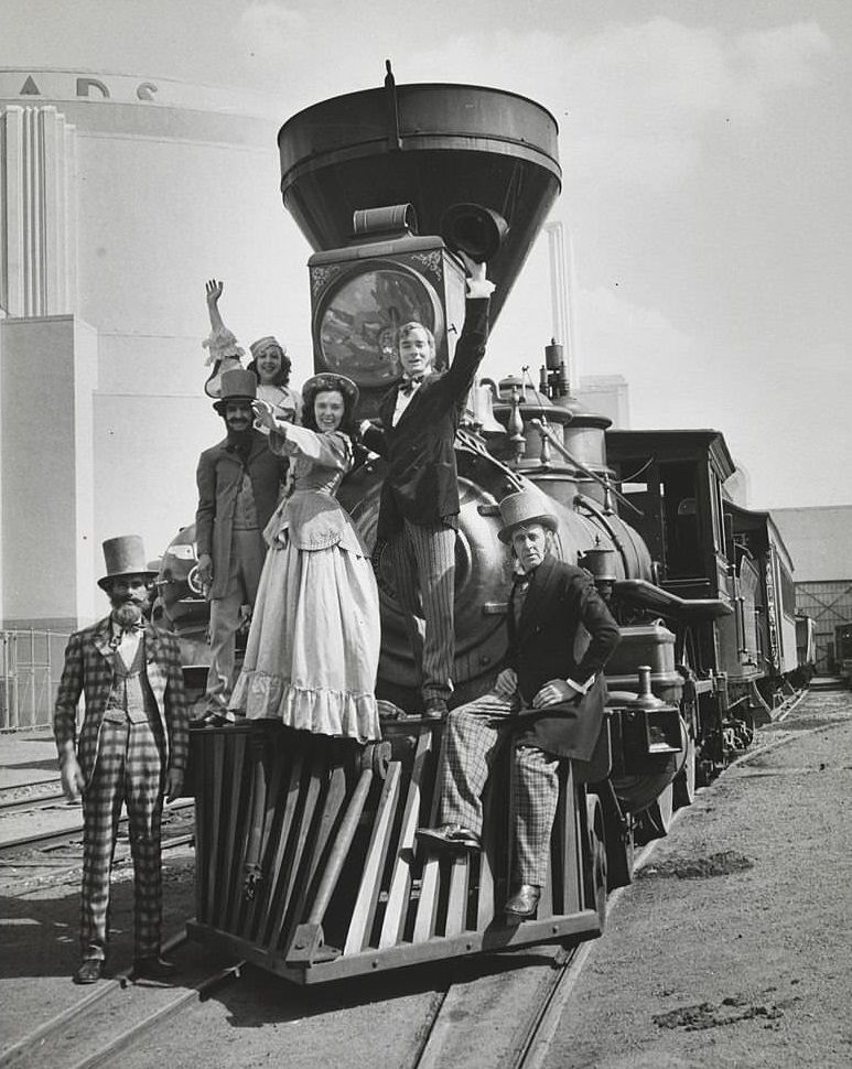 View showing a group of actors in various costumes posed on the front of a steam engine.