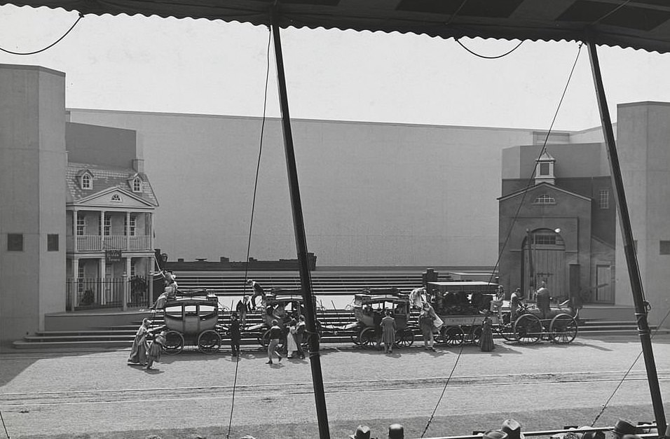 View showing an outdoor performance featuring actors in costume (top hats, full dresses, ect.) on a set featuring a small steam engine pulling a covered passenger car and several stagecoaches.