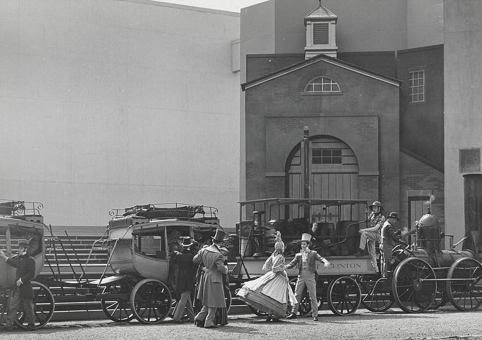 An outdoor performance featuring actors in costume (top hats, full dresses, ect.) on a set featuring a small steam engine pulling a covered passenger car and several stagecoaches.
