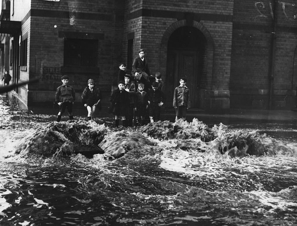 Children watch as a torrent of water pours out of drains in a Glasgow street during severe floods throughout Scotland, 1964