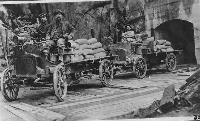 Three men riding in an automobile and trailers for hauling cement, 1906