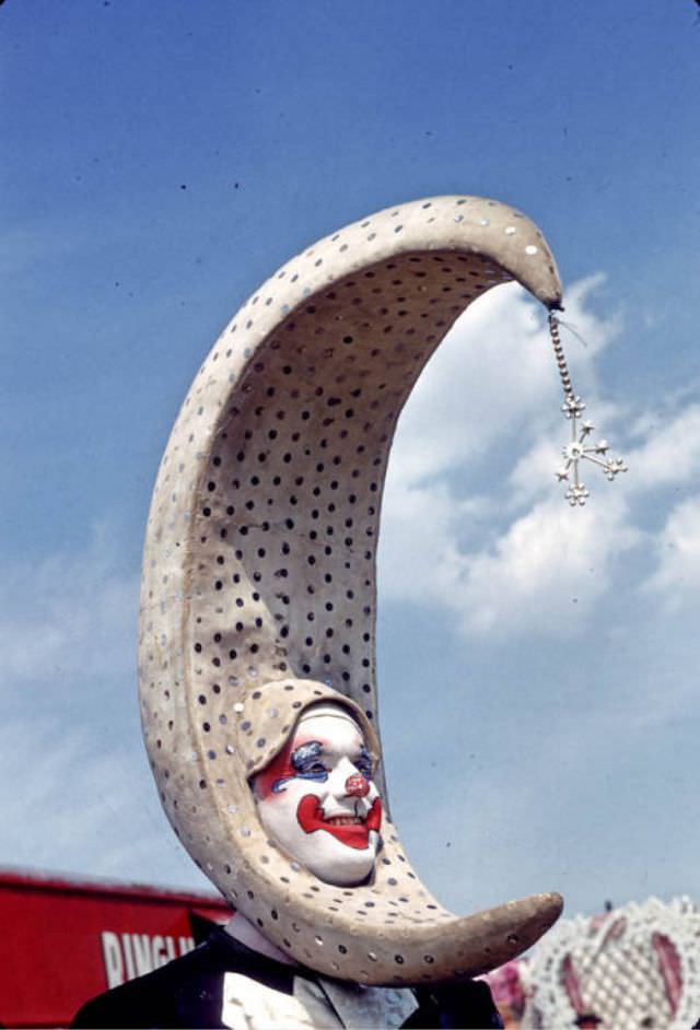A clown with a large crescent head piece in Holidays spec wardrobe.