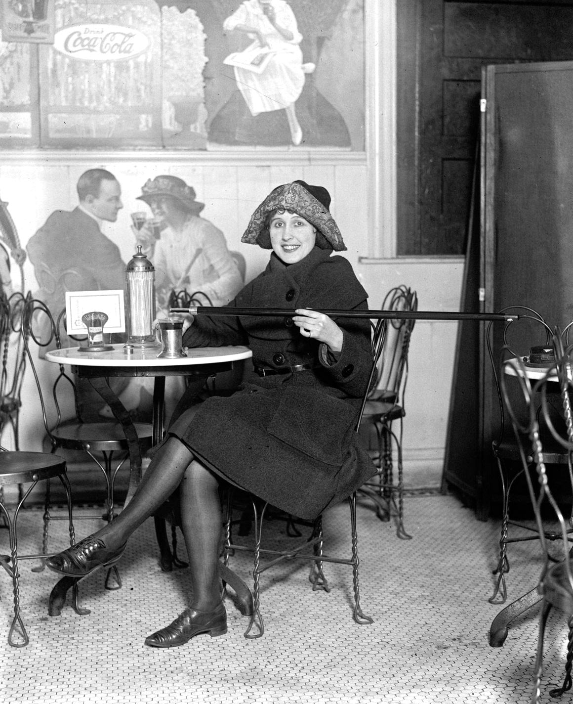 A woman in Cloche Hat, seated at a soda fountain table, as she pours alcohol from a cane into a cup during Prohibition, 1922