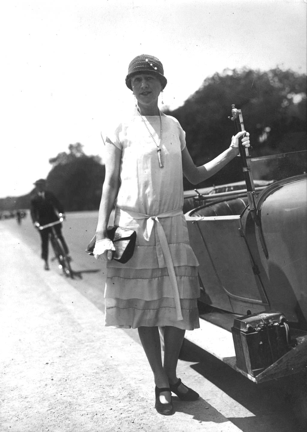 A model wearing a pleated summer dress and cloche hat whilst leaning on a car, 1925
