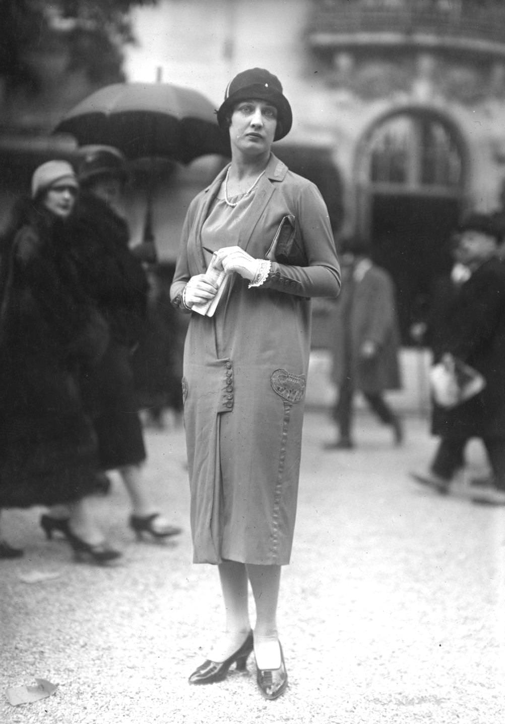 A model stands in the rain, wearing a linen tunic coat with buttoned panel at hip height, and a cloche hat with small brim, 1925