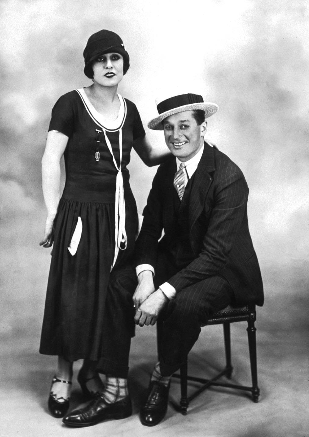 Yvonne Valle standing beside French singer and actor Maurice Chevalier as he sits on a chair and smiles. Valle wears a cloche hat with a day dress; Chevalier wears a three-piece pinstriped suit and a straw boater, 1921
