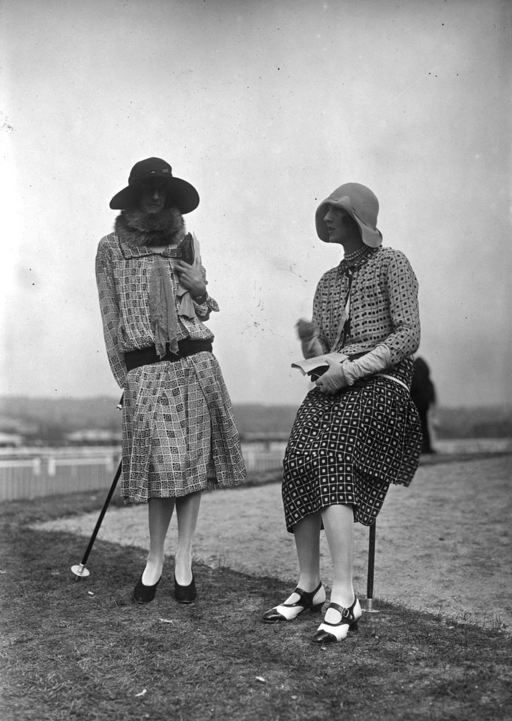 Two racegoers wearing long-waisted patterned dresses with panels One has two-tone one-bar shoes and her cloche hat has a wide turned-down brim at left-hand side, 1925