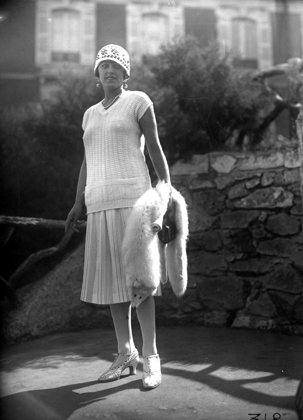 Cap sleeved v-necked jumper worn with a pleated skirt, knitted cloche hat and t-bar high-heeled shoes. Accessories include a white fox fur,short single-strand pearl necklace, and drop earrings, 1925