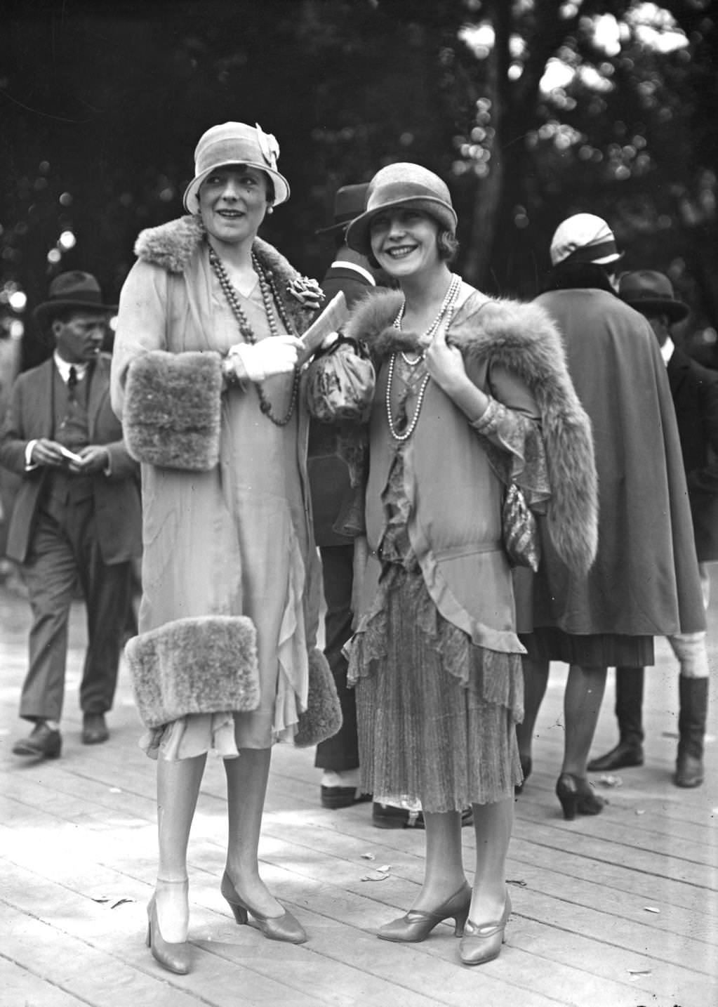 Two outfits designed by Jenny. One is a light coat with wide fur trim at hem cuffs and collar worn with a cloche hat with a small brim, a double rope of beads and an anklet chain, 1925