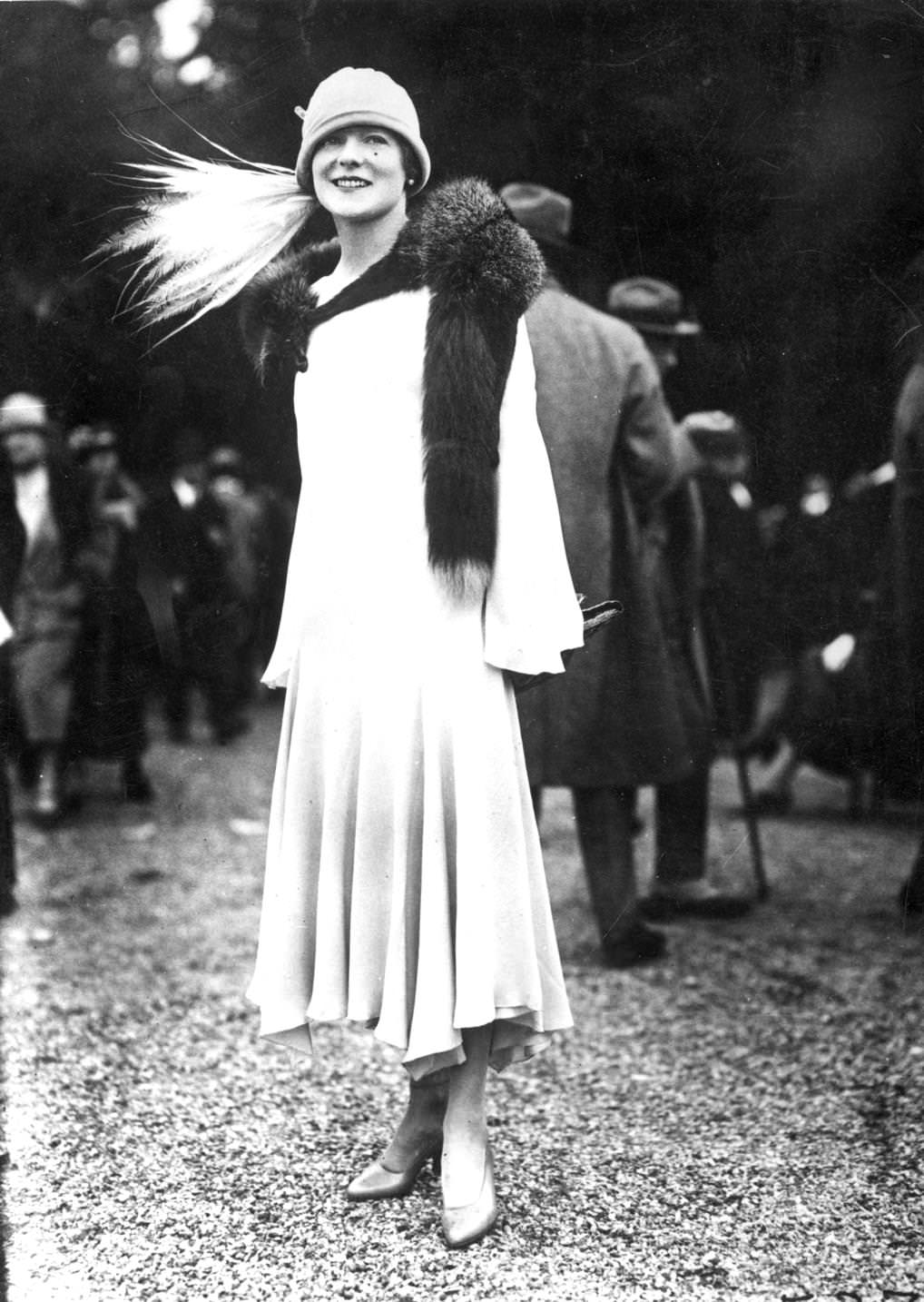 An elegant evening dress with a tapering uneven hemline and sleeves. A fur stole and hat with an array of long white feathers complete the look, 1925