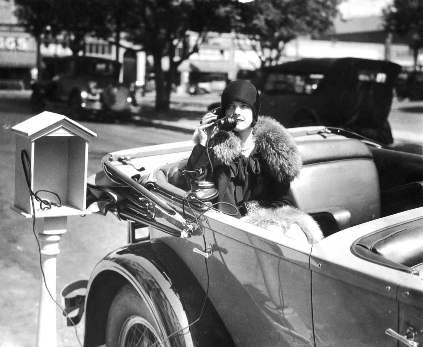 A woman sits in an automobile while talking on the telephone from a phone service stand at the Hotel Constance in Pasadena, California. She is wearing a cloche hat and a fur-trimmed coat, 1925