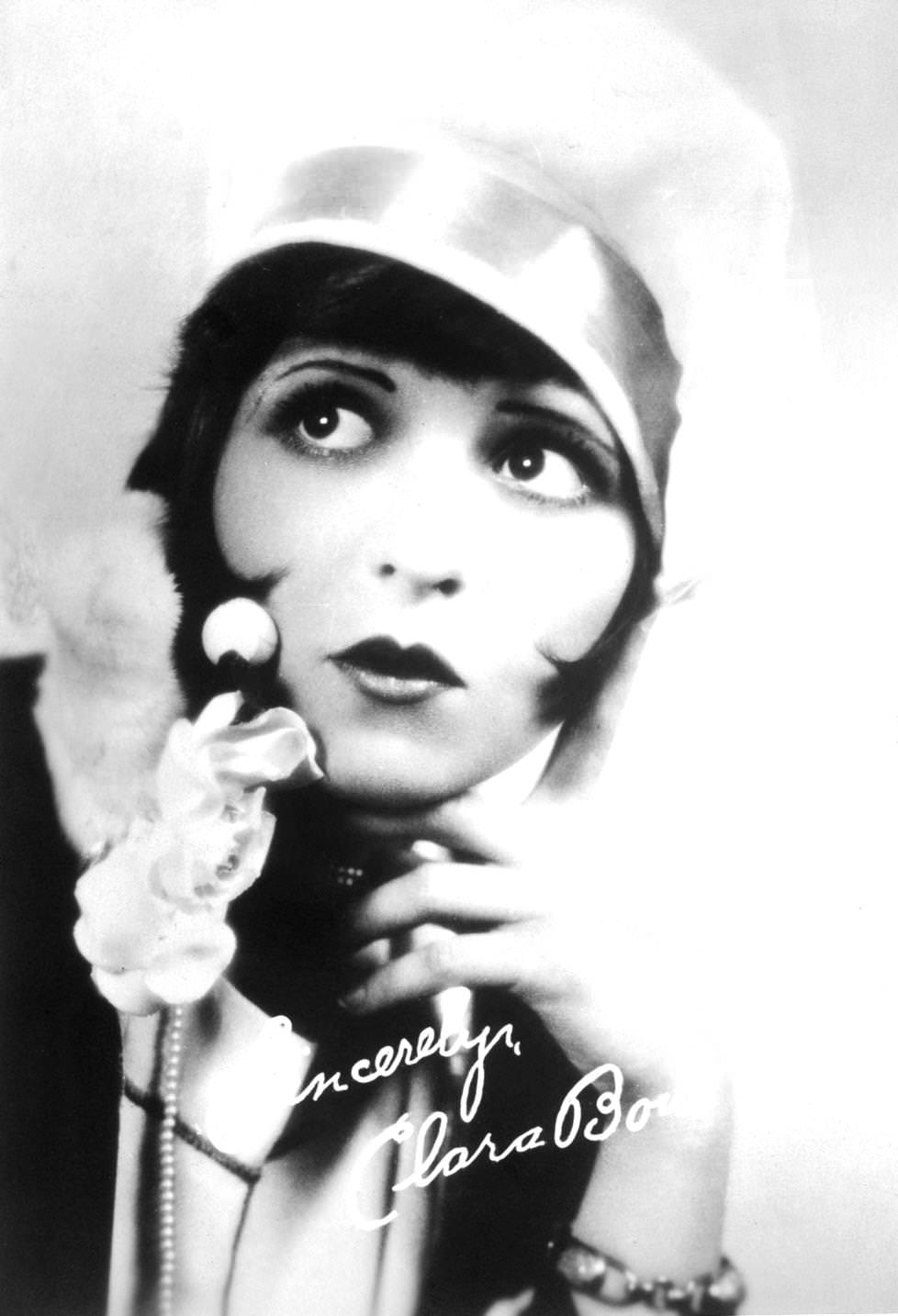 Autographed studio portrait of American actor Clara Bow wearing a cloche hat and a bobbed hairstyle while holding her hand to her face and pursing her lips, 1925