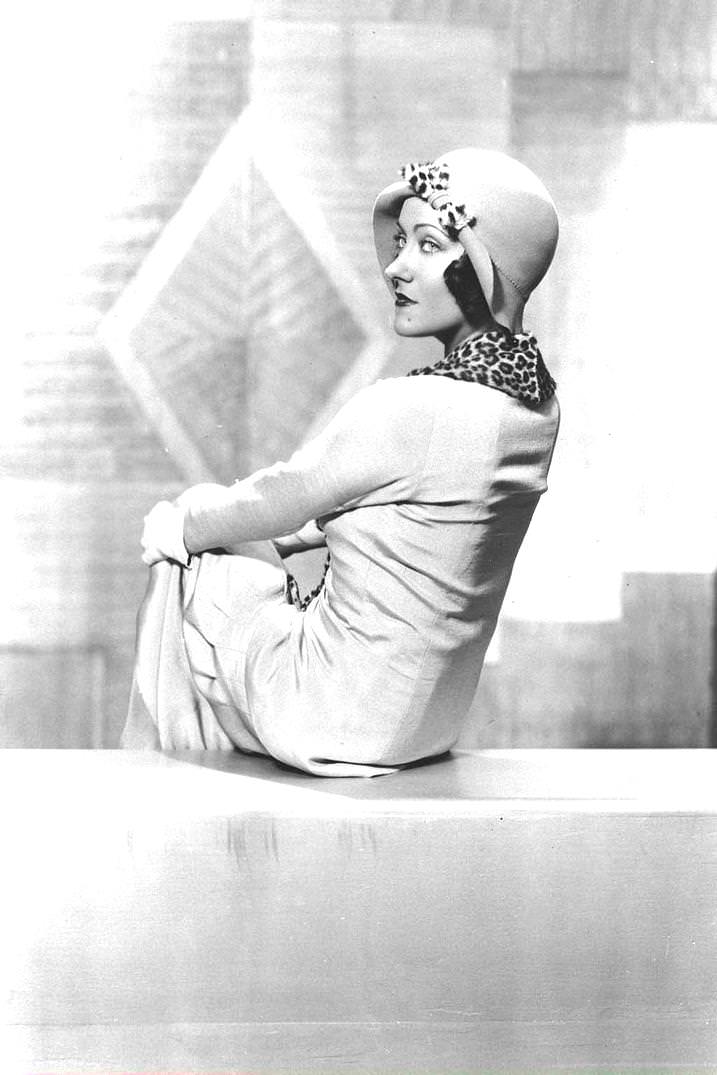 American film actor and producer Gloria Swanson sits with her knees crossed and her back to the camera as she looks over her shoulder. She wears white gloves, a cloche hat with a leopard print bow, and a matching scarf, 1926