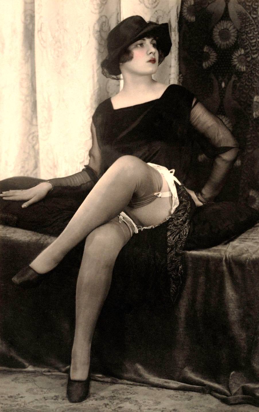 A woman seated on a divan, she wears a square necked black velvet dress with sheer chiffon sleeves and a black cloche hat, 1920