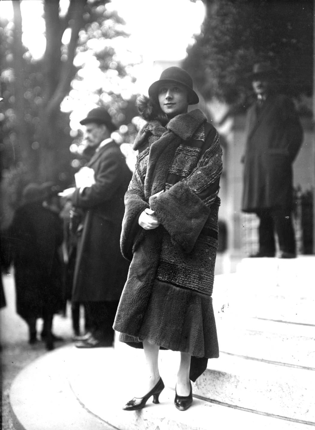 A brocade coat trimmed with lamb designed by Drecoll, worn with plain high-cut court shoes and a cloche hat with a brim, 1924