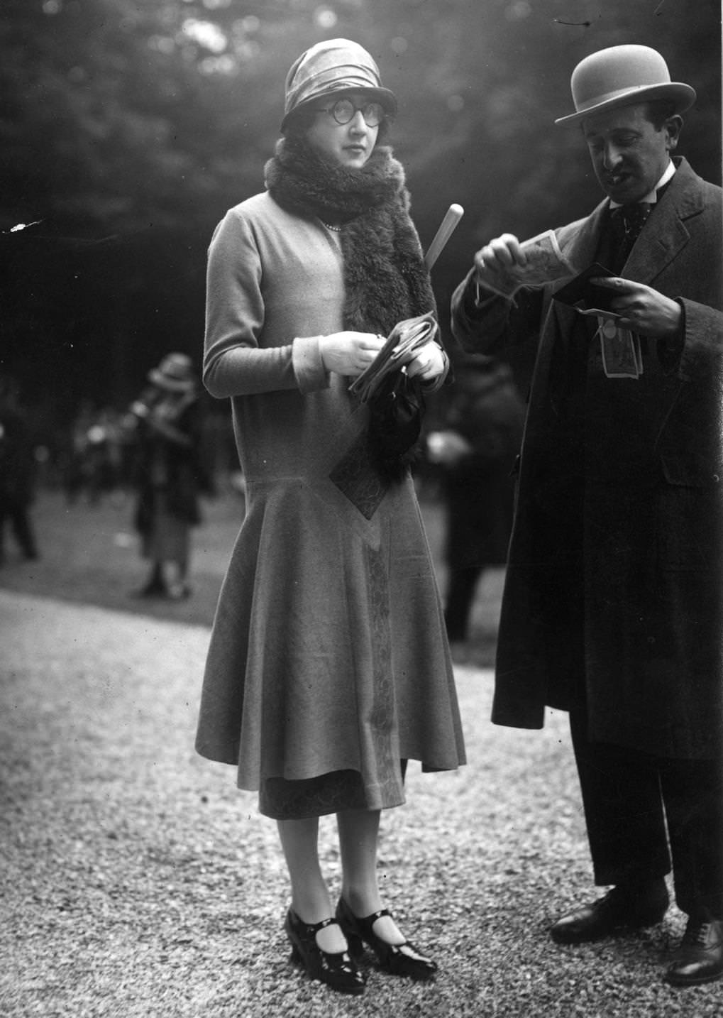 A woman wears a plain long waisted seven eighths coat worn with a cloche hat and a fur scarf. Her male friend puts away some large banknotes, 1924