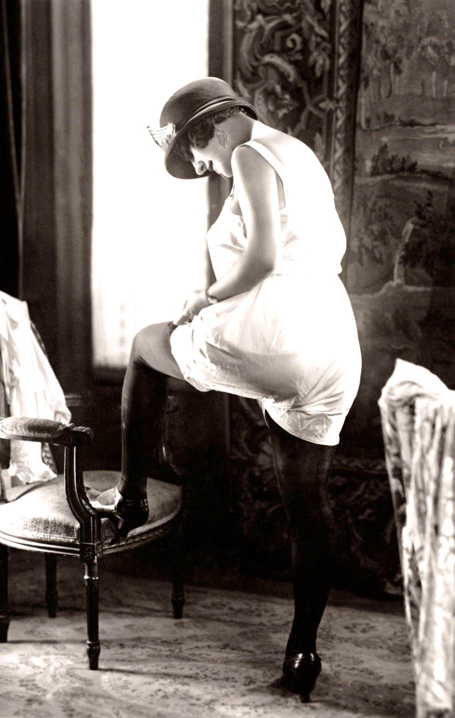 A young woman fastening her stockings and wearing a loose fitting 'step-in' chemise undergarment and a cloche style hat, Paris, 1920.