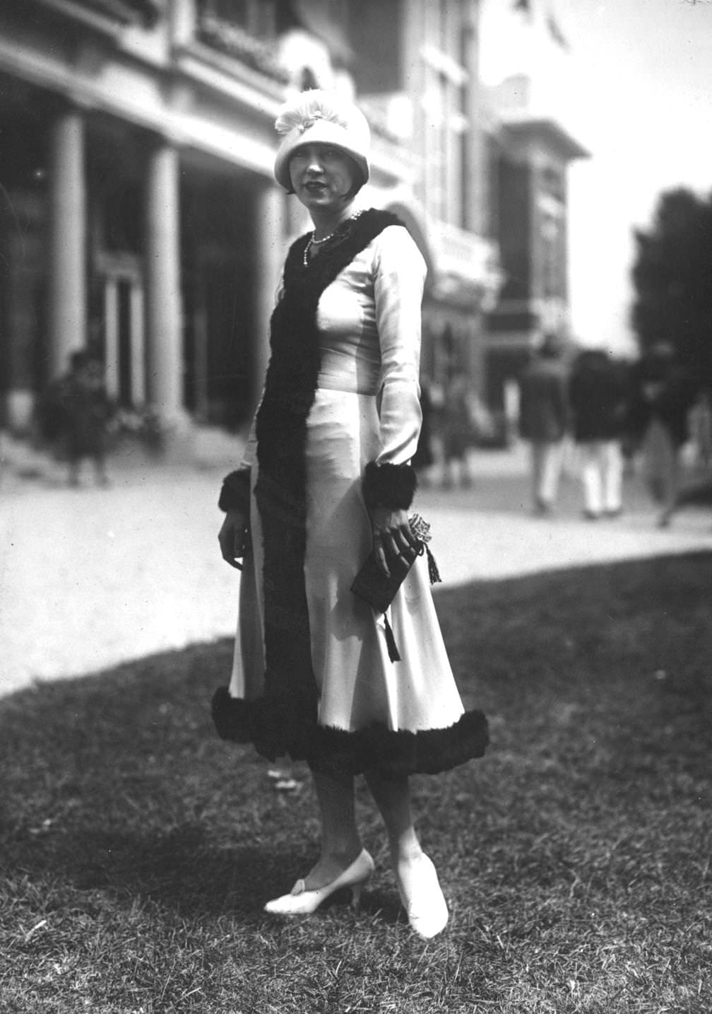 The model wears a fur trimmed coat with cloche hat decorated with a small plume of feathers at the brim. She carries a small clutchbag fringed with tassels, 1925