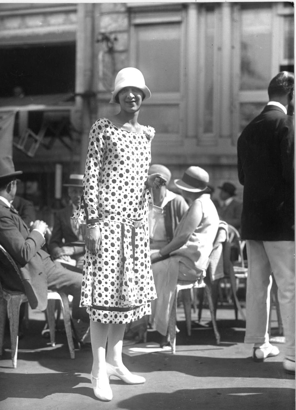 Long-sleeved spotted dress, boat neckline and dropped waist with pleats over the hips. Cloche hat with turned back brim, white stockings and one-bar shoes, 1925