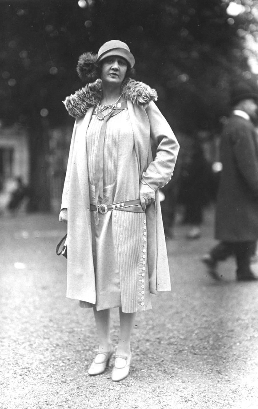 Low waisted calf length dress with buttoning side panels along one side. Coat with fur collar, and cloche hat with fur pom-pom, 1925