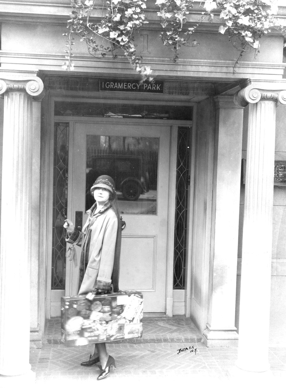 Journalist Mabel Herbert Urner, in cloche hat, standing on pillared stoop of her 1 Gramercy Park house, holding suitcase covered with travel stickers, New York, 1922.