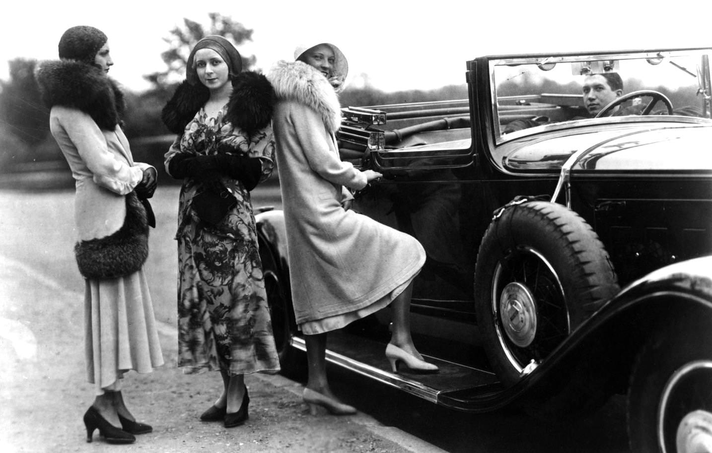 Women standing by a convertible car, wearing fur lined coats and cloche hats, 1920