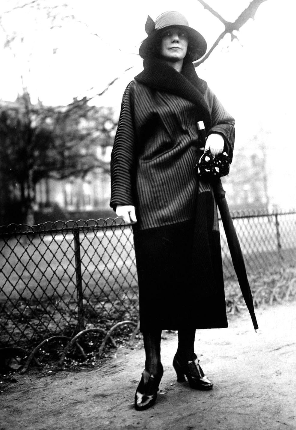 Fashion model wearing a short wrapover coat with fur collar, brimmed cloche hat, straight skirt and carries an umbrella, 1920s