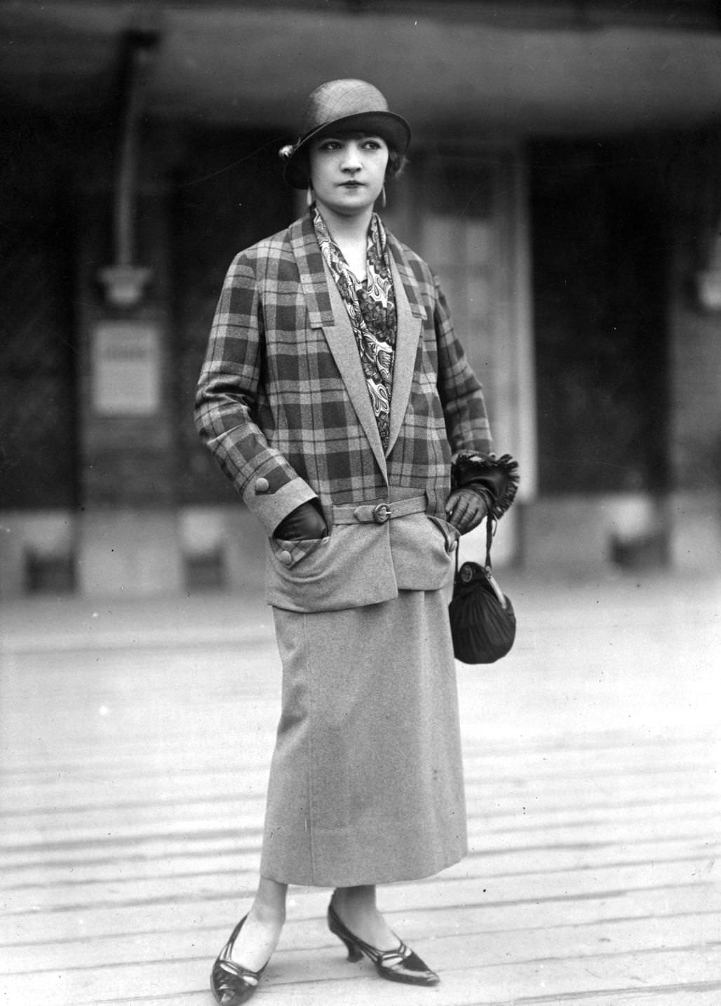 V-necked suit by Felix Dupouy. Check jacket has dropped waist worn over a plain skirt. Hat is a small brimmed cloche, small pouch shaped handbag is carried over the wrist, 1924